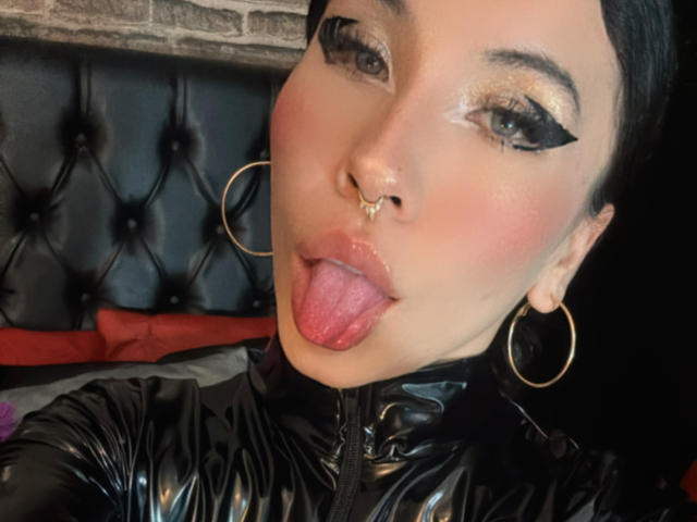 SofiBrandford has black eyeliner, gold glitter, and a nose ring. She's sticking out her tongue, with pink lips, and wearing a black leather zip-up. Large gold hoop earrings.