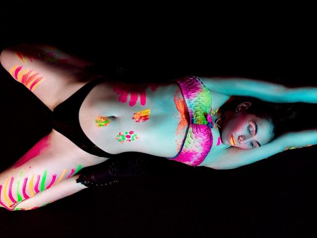 AngyWahls is lying down with arms above her head, covered in neon paint on her tits, ass, and belly, wearing black panties, and black boots, in a dark room.