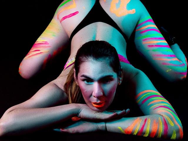 AngyWahls is on her back with legs up, black thong, neon paint on her body, face, and arms, looking at the camera with a serious expression, black background.