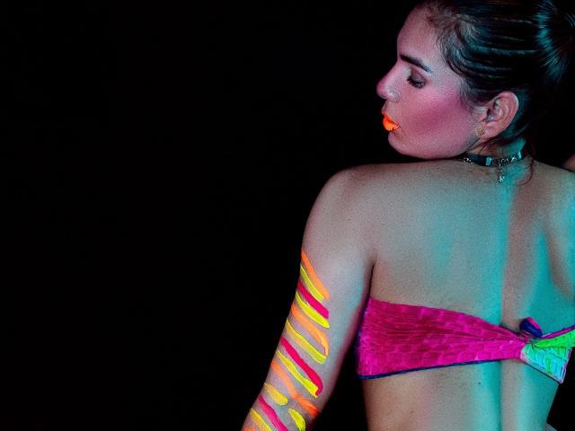 AngyWahls has neon orange and yellow swirls on her right arm, wearing a pink strapless top, and has orange glow-in-the-dark lipstick. She's looking down, with dark hair.