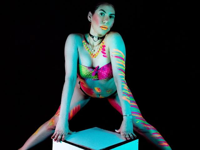 AngyWahls with neon paint on her tits, ass, and legs, wearing a colorful bra and choker, crouching on a glowing box, against a black background.