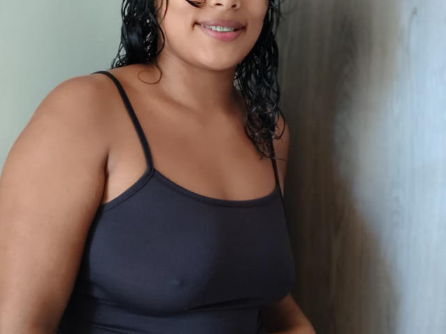 VelvetAndNoiir with wet black hair, smiling, wearing a tight black spaghetti-strap top, showing her medium-sized tits and tan skin.