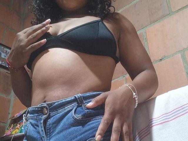 VelvetAndNoiir with dark skin, curly hair, black bra, and blue jeans, showing her tits and belly, hand on hip, in a bathroom, wearing bracelets.