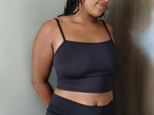 VelvetAndNoiir has tan skin, wet black hair, small dark nipples, and wears a tight black sports bra, showing her boobs and a flat stomach, with hands behind her.