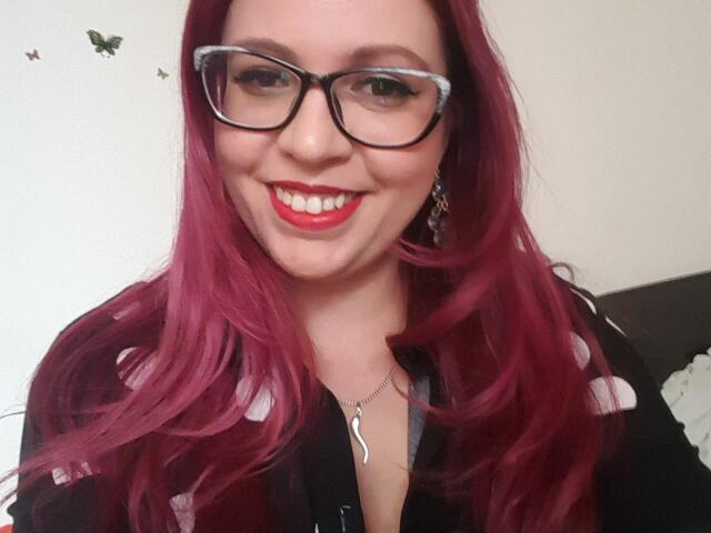 LadyHellena with red hair, black glasses, red lipstick, smiling, wearing a black shirt with white polka dots, necklace with a crescent moon pendant.
