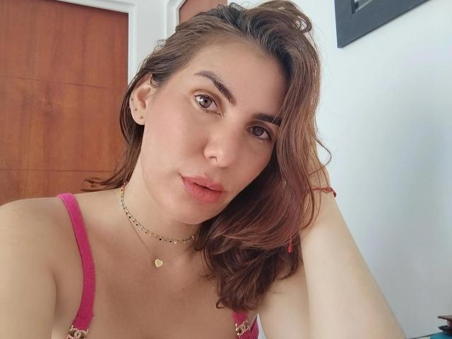 AngyWahls has brown eyes, wavy brown hair, pink bra, gold necklace, and pink lips. Her tits are medium, and she's looking at the camera with a slight smile.