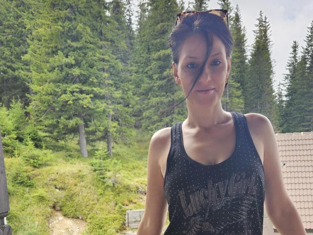 JoliDanielle with small tits, wearing a black sparkly tank top, has brown hair, sunglasses on head, and a slight smile, standing in a green forested area.