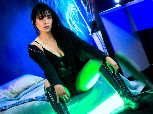 MelaniePower with black hair, wearing a black dress and lace-up boots, sitting on a bed with green and blue lights, showing her tits and looking at the camera.