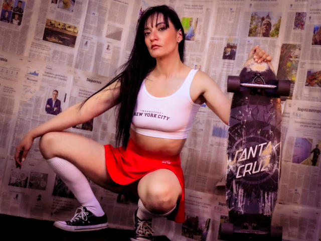 MelaniePower, squatting, wearing white crop top, red skirt, black sneakers, white knee socks, holding black and white "Tana Cru" skateboard, long black hair, pale skin, small tits, confident.