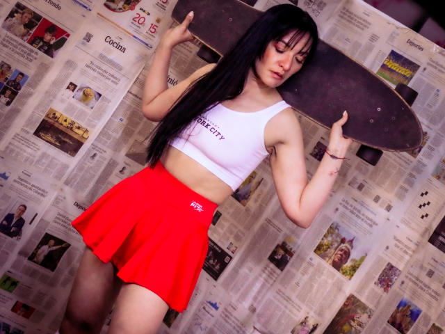 MelaniePower with long black hair, white crop top, red skirt, holding a skateboard, showing a slim, fit body, small tits, and a confident, sexy pose.
