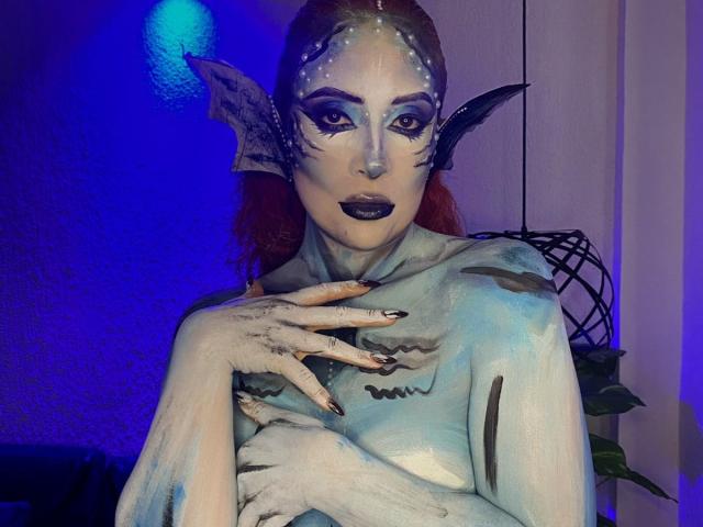 Sahrataylorr with painted blue-green skin, dark makeup, fish-like ears, and black nails, touching chest. Red hair, underwater theme, standing, hand on chest, blue and purple lighting.