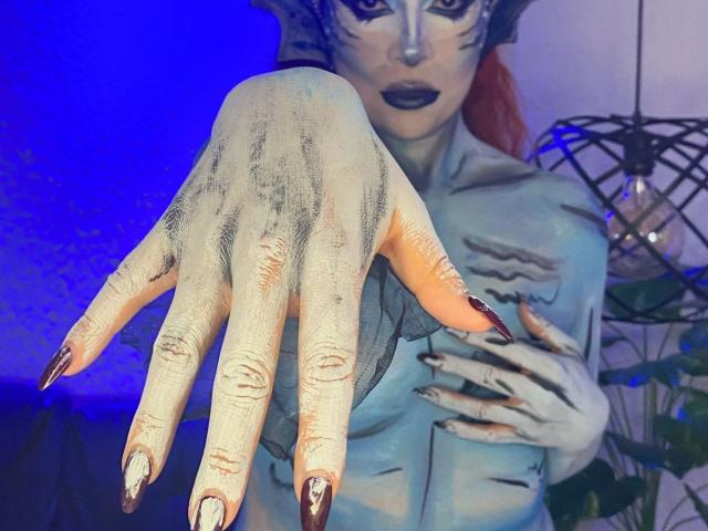 Sahrataylorr's pale, grimy hand with long, dark, pointed nails reaches forward, painted to resemble a zombie's. Sahrataylorr's face is painted with black markings, wearing dark lipstick.