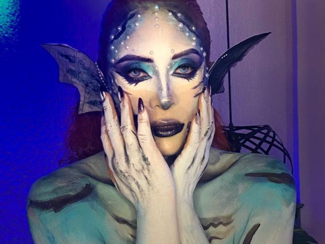 Sahrataylorr with mermaid makeup, blue-green skin, black lipstick, fin ears, and hands on face, looking intense with glowing eyes, in a purple-blue lit room.