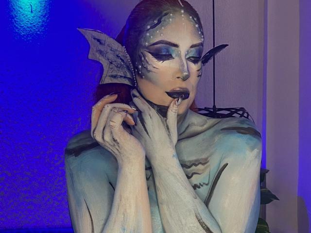 Sahrataylorr is a painted mermaid with grayish-blue skin, dark makeup, and a fin-like ear, holding her finger to her mouth, looking sultry with blue lighting.