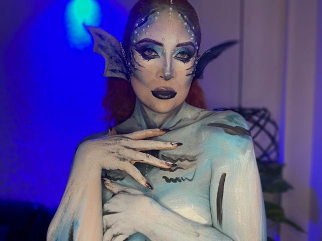 Sahrataylorr, a pale, blue-painted mermaid with red hair, black lipstick, and fish-like ears, covering her tits with gray, webbed hands, against a blue-lit background.