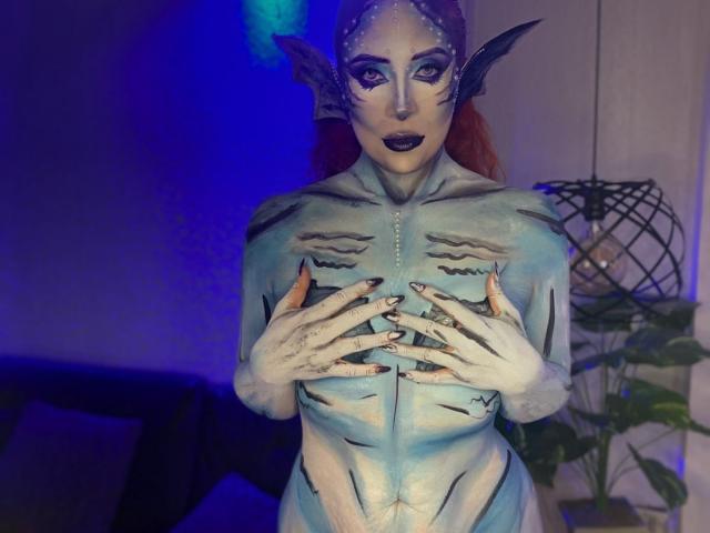Sahrataylorr with painted mermaid-like face, blue body paint, and fin ears, covering her tits with white-painted hands, in dim blue-lit room, wearing dark lipstick.