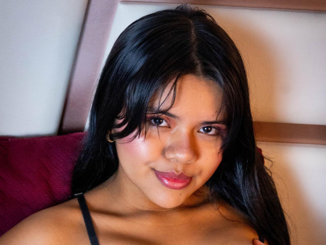 AmbarRichard has long black hair, brown eyes, pink lipstick, and a cute smile. She's wearing a black spaghetti strap top, and her small tits are slightly visible.