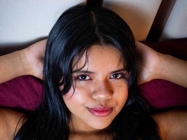 AmbarRichard has dark black hair, brown eyes, and a slight smile. She's lying on a red pillow with her hands behind her head, showing her cute, small nose.