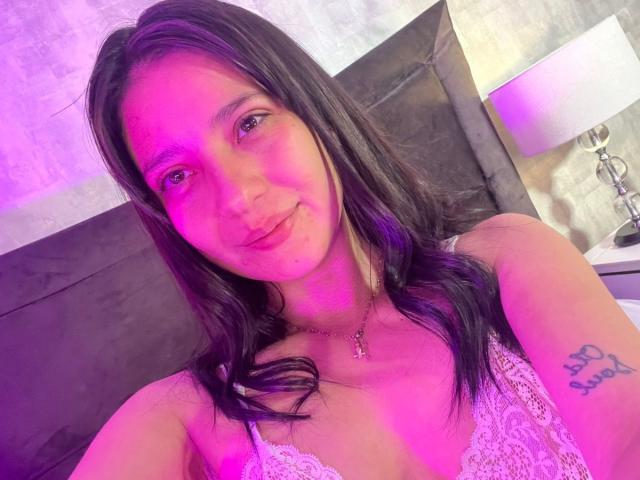 CattsyCathy with long black hair, pink-tinged skin, and a lace top, showing a tattoo on her right arm, smiling with pink-lit face, in a bedroom.