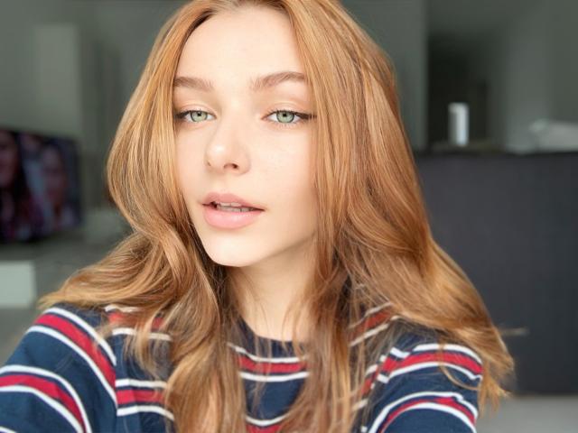 AmberCrazy has light green eyes, a cute nose, and full pink lips. She's wearing a striped shirt with red, white, and blue lines, and has wavy, red hair.
