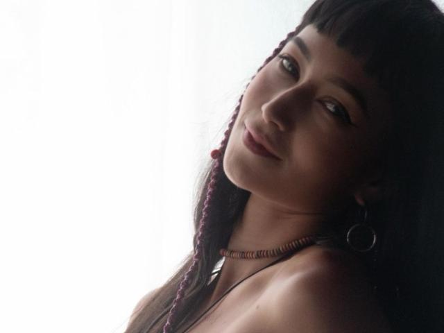 NofaceNice has dark hair in braids, wearing hoop earrings, looking over her shoulder with a slight smile, her bare shoulders visible, soft light on her face.