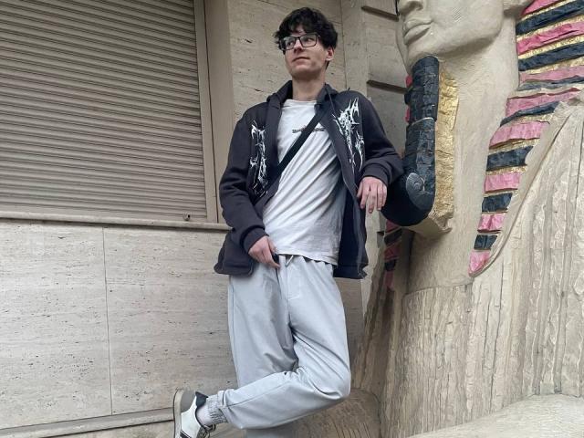 Denrich with curly black hair, glasses, black hoodie, white tee, grey pants, black and white sneakers, standing with hand on skateboard, leaning against beige wall with stone texture.