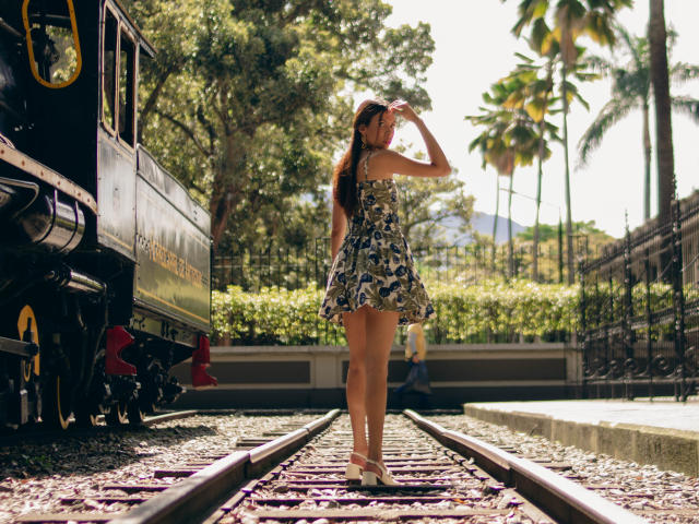 ElevenSkys in a floral dress, standing on railroad tracks, back to camera, hand on head, black train on left, palm trees, sunny, wearing white heels, looking over shoulder.