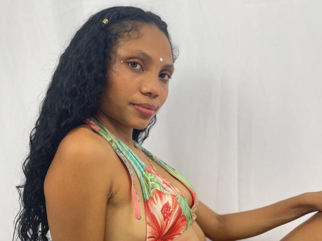 LatinDeepHard with dark curly hair, small nose dots, green and red floral bikini, brown skin, sitting, white background, looking at camera, subtle smile, small gold hair clip.