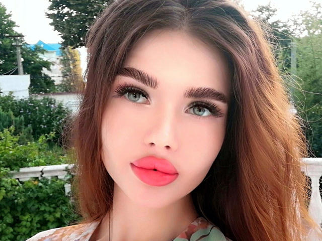 KatrinaBonita-hot has light green eyes, thick brown eyebrows, long eyelashes, pink lipstick, and wavy brown hair. Her face is centered in the image.