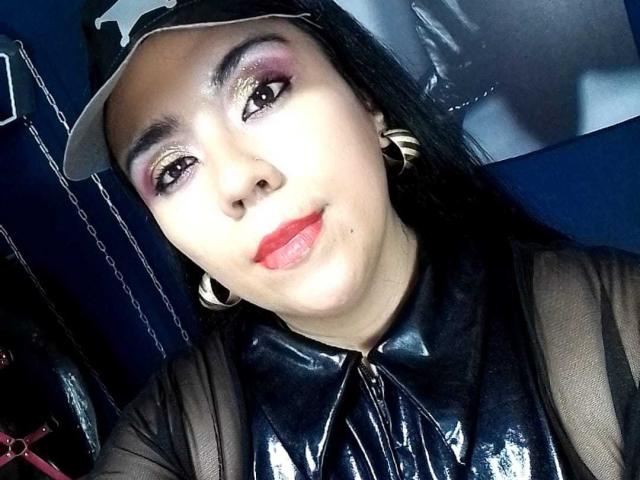 LexyDom with shiny purple eyeshadow, red lipstick, black latex shirt, and gold hoop earrings, wearing a white cap, has a cute smile and a slightly chubby face.