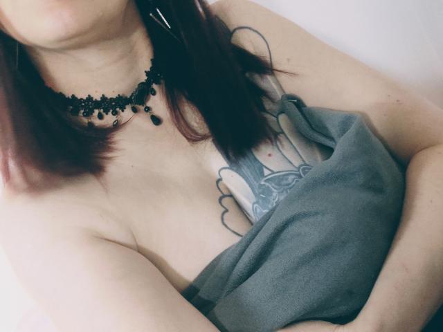 CatalinaMonyque's tits are covered by a grey towel, showing a tattoo of a cloud and a face between her breasts. She has purple-dyed hair and a black bead necklace.