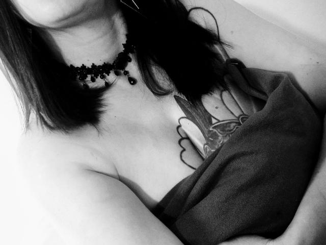 CatalinaMonyque com colar preto, cabelo curto e escuro, top sem alças, tatuagem de uma mão com uma borboleta no peito, braços cruzados, foto em tons de cinza, decote visível, textura suave..