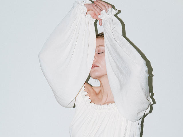 Eolanta Marina, con los ojos cerrados y los brazos levantados, vestía un vestido blanco con un hombro descubierto y mangas largas, con las manos agarrando la tela. Sus pechos se veían ligeramente debajo del vestido..