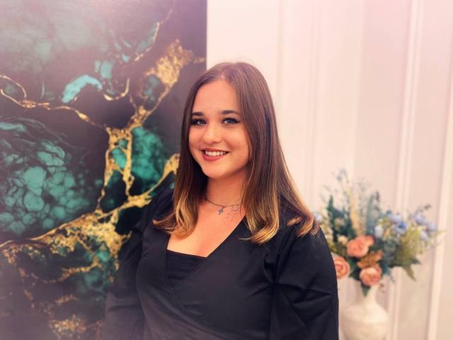 MiaDaweryopi has shoulder-length brown hair, black V-neck shirt, and is smiling with red lipstick. She's wearing a necklace, standing in a room with floral decor.