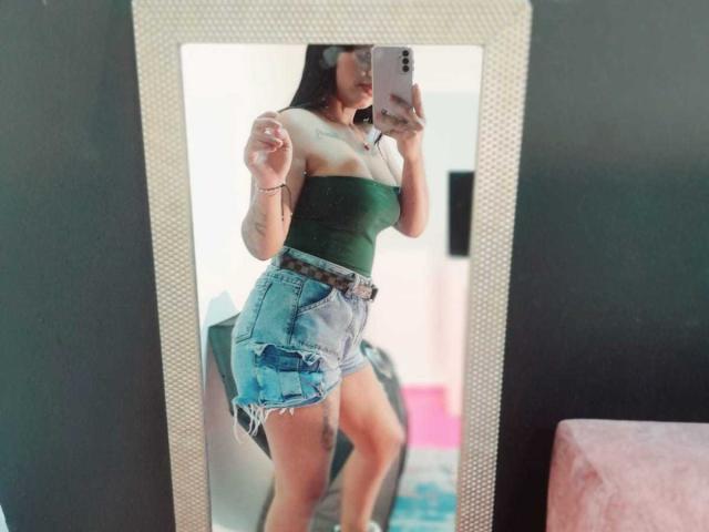 GiaAttum macht ein Selfie vor einem Spiegel und zeigt ihre Brüste in einem grünen, trägerlosen Top und zerrissenen, blauen Jeansshorts, mit einem braunen Gürtel und einem Tattoo an ihrem linken Oberschenkel..