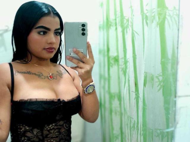 GiaAttum with black lace top, showing her big tits, holding a phone, green bamboo curtain, long black hair, red lipstick, tattoos, red heart necklace, and a wristwatch.