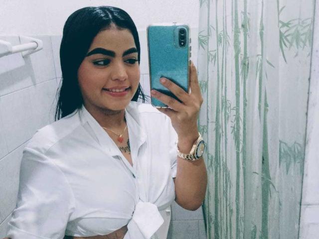 GiaAttum, con cabello oscuro, camisa blanca anudada, sosteniendo un teléfono azul, sonriendo, usando un reloj dorado, un collar y un colgante rojo, tomando una selfie en el baño con una cortina de bambú..