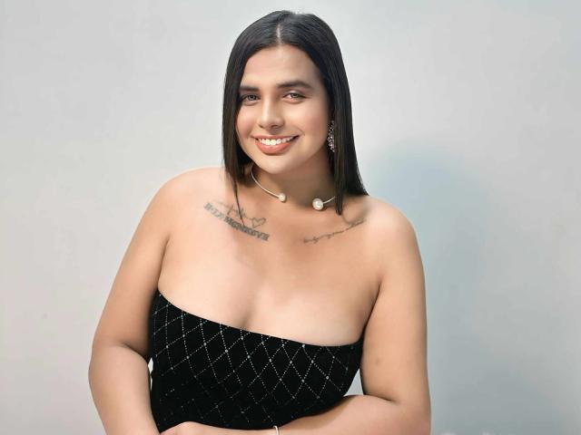 HotSelenaTrans with straight black hair, smiling, wearing a black strapless dress with diamond patterns, small tits, tattoos on chest, white shell necklace, and silver earrings.