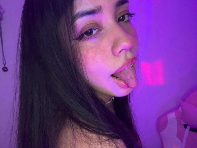 FlameAmber69 has long dark hair, brown eyes, and freckles, sticking out her pink tongue with purple-lit face, wearing black eyeliner and a choker.