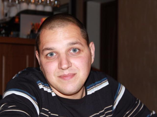 MichaelSexos, chubby-faced, bald with short hair, blue eyes, wearing a black and white striped shirt, smiling slightly, sitting in a dimly lit room.