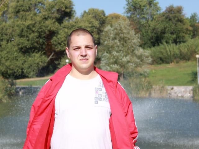 MichaelSexos has a chubby face, short buzzed hair, wearing a red jacket over a white shirt, standing in front of a lake with trees in the background.