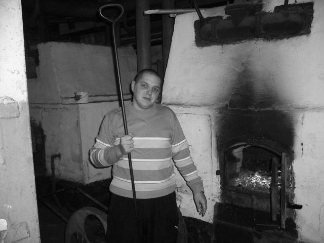 MichaelSexos, a chubby dude with a shaved head, wears a striped sweater and holds a shovel, standing by a blackened oven in a dimly lit, grimy basement.