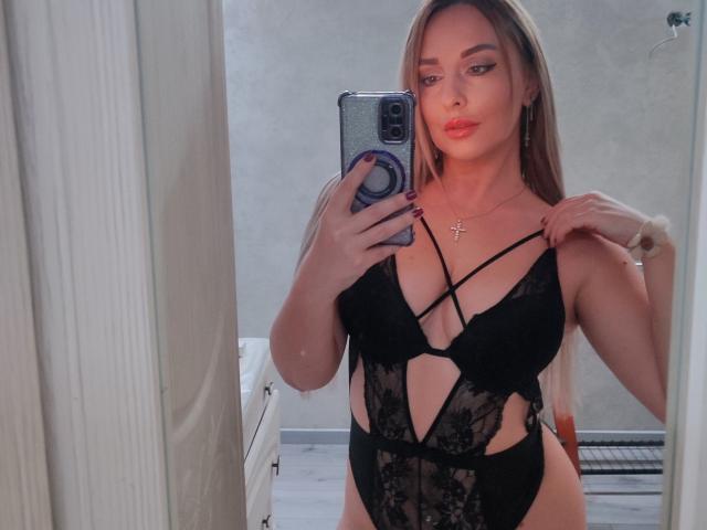 SoNakeDream in black lace lingerie with cutout sides, holding phone in mirror, showing her tits, with long blonde hair, red lipstick, and a cross necklace.