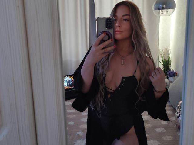 SoNakeDream with long blonde wavy hair, black lace-up top revealing her boobs, holding phone in mirror selfie, wearing black jacket, cross necklace, and red nails.