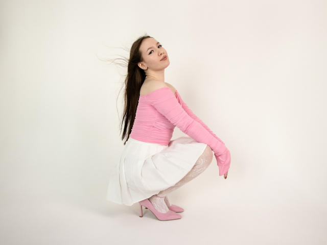DanielleSweetty, squatting, wearing pink off-shoulder top, white skirt, pink high heels, and white tights, with long brown hair, pink lipstick, and a slight smile.