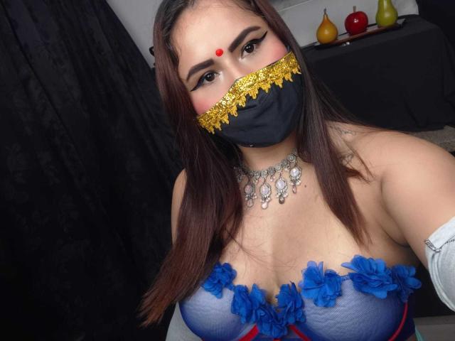Dalia Dubey, met bruin haar, draagt een zwart masker met gouden kant, een rode stip op haar voorhoofd, een blauwe jurk met bloemen, waarbij haar grote borsten zichtbaar zijn, een zilveren ketting, een witte arm, een zwart gordijn..