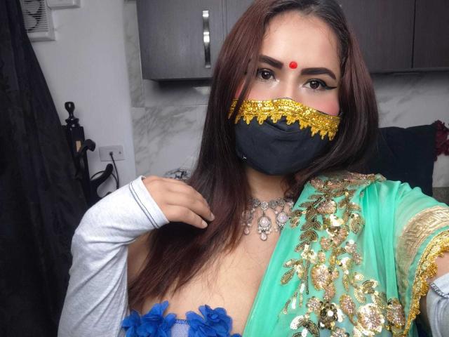 Dalia Dubey, met lang bruin haar, draagt een groene sari met gouden borduursel, een zwart masker met gouden rand, een rode stip op haar voorhoofd, een blauwe bloem in de decolleté en een zilveren ketting..