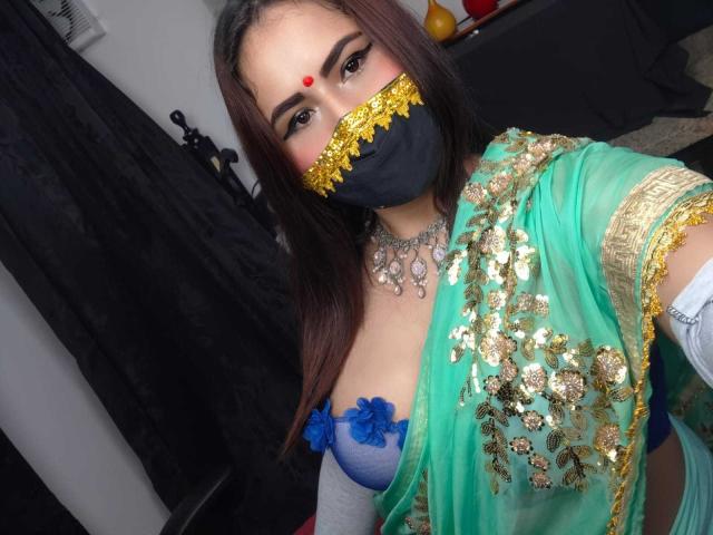 DaliaDubey with dark hair, wearing a green and gold traditional outfit, black mask with gold trim, red bindi, blue bra with blue flowers, showing her tits, and a necklace.