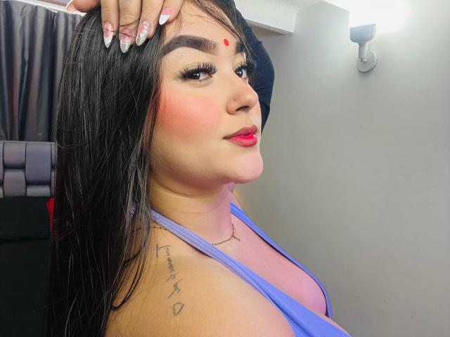 AnaShalimarr, con cabello oscuro, un punto rojo en la frente, rubor rosado, lápiz labial rojo, blusa azul, tatuaje en el hombro derecho, mano en la cabeza, uñas largas, mirando por encima del hombro, sonrisa suave..