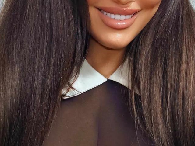 O rosto de LiaaEve, com lábios rosados e brilhantes, mostrando os dentes superiores, vestindo uma blusa preta transparente com uma gola branca, e cabelos castanhos longos e lisos em ambos os lados. Os seios estão visíveis através da blusa..